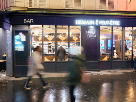 La facade du café "Demain Peut-être" illuminée de l'intérieur, des passants flous passent devant.